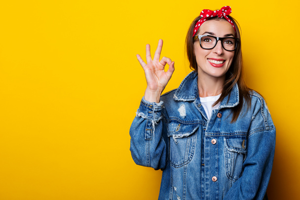Smiling young woman in hair band, in denim jacket shows ok gesture, okay on yellow background - Fotó, kép
