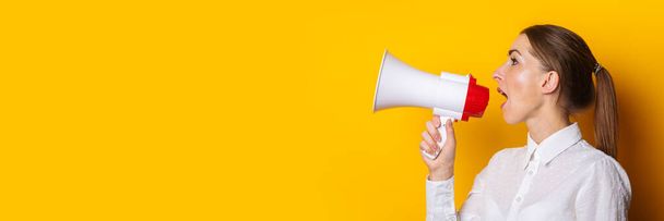 Young woman in a white shirt shouts into a megaphone on a yellow background. Concept for hiring, listing, help wanted. Banner. - Foto, Imagem