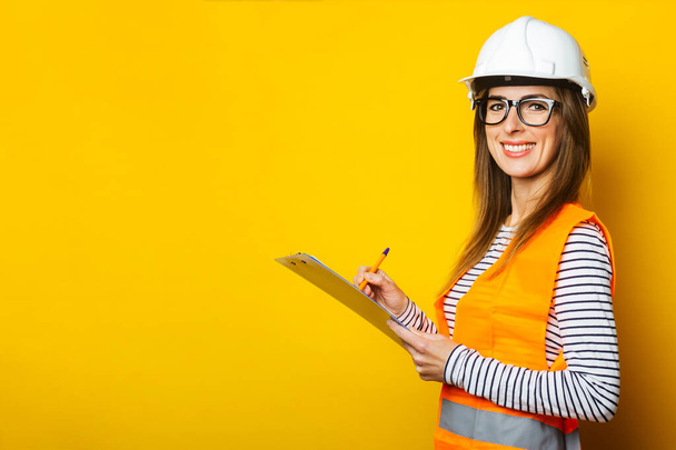 Young woman in a vest and hard hat holds a clipboard and takes notes on a yellow background. Concept for construction, new building, renovation. Banner. - Photo, Image