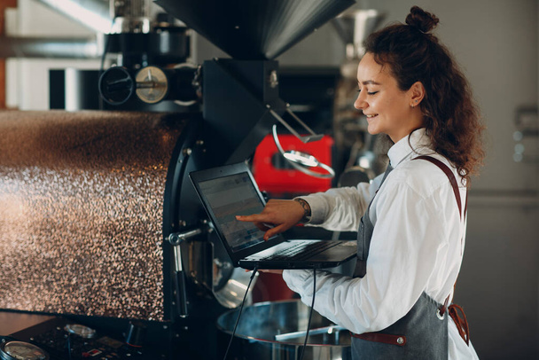 Coffee roaster machine and barista woman with laptop at coffee roasting process - Foto, afbeelding