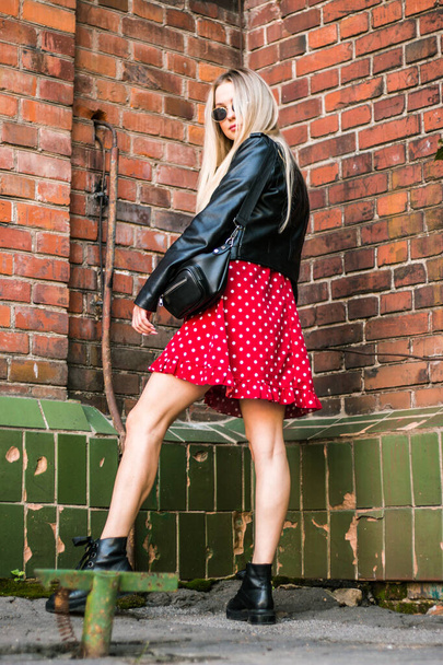 urban street portrait of young beautiful stylish woman in red dress and black leather jacket. Standing near an old brick wall, natural evening light - Φωτογραφία, εικόνα