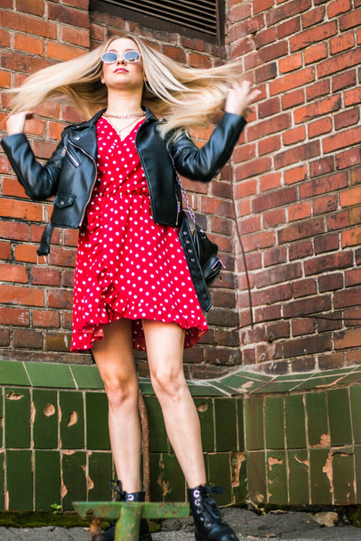 stedelijk straatportret van jonge mooie stijlvolle vrouw in rode jurk en zwarte leren jas. Staande in de buurt van een oude bakstenen muur, natuurlijk avondlicht - Foto, afbeelding