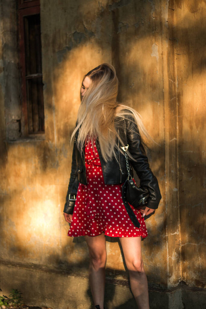 urban street portrait of young beautiful stylish woman in red dress and black leather jacket. Standing near old grunge wall, natural evening light, sunset - Foto, Imagen