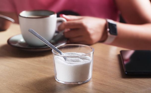 Woman with clock and phone holding a cup of coffee with milk and next to sugar substitute in a cafe - 写真・画像