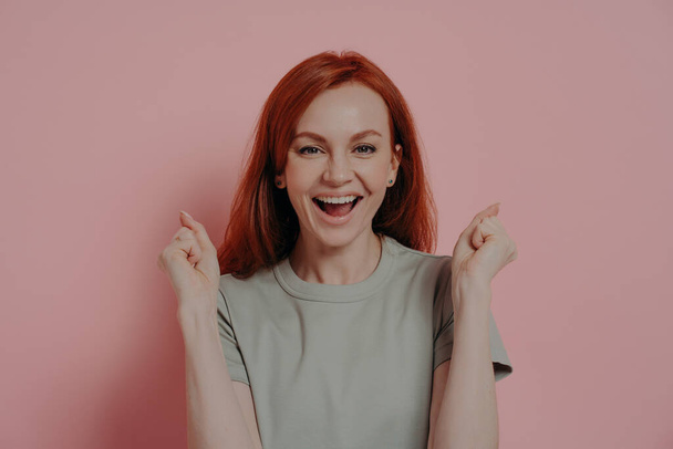Young overjoyed redhead woman raising hands up with clenched fists, doing champion gesture - Foto, afbeelding
