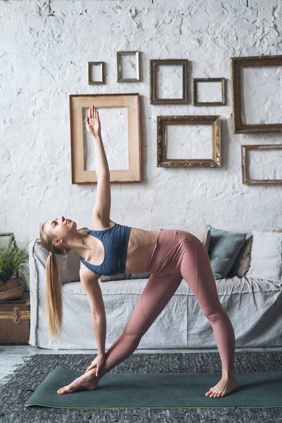 Vertikale Aufnahme einer schönen, fitten Frau in guter Verfassung, die Morgengymnastik macht, während sie in Yoga Asana Utthita Trikonasana im Wohnzimmer zu Hause steht, eine sportliche Frau in aktiver Kleidung, die drinnen trainiert - Foto, Bild