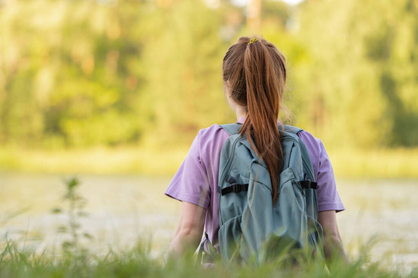 Punapää tyttö istuu nurmikolla lähellä vettä kauniissa kesämetsässä. Nuori nainen matkustaa reppu - Valokuva, kuva