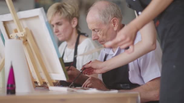 Nuori naisopettaja auttaa vanhaa eläkeläistä oppimaan maalaamaan kuvan. Vanhempi mies oppii piirtämään - Materiaali, video