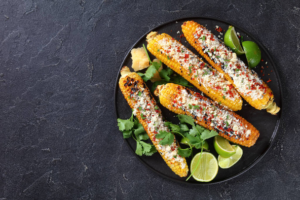 Elote on a black plate, Grilled Mexican Street Corn, charred cobs are slathered in sour cream based sauce, seasoned with chili powder and sprinkled with cheese, cilantro, flat lay, free space - Foto, Imagen
