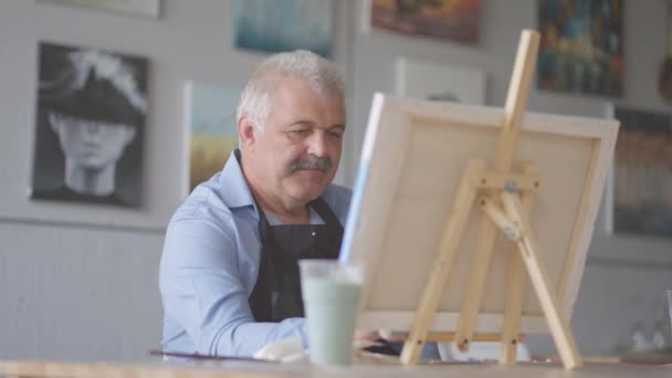 Een oudere man in een schort schildert een schilderij met een borstel terwijl hij aan tafel zit - Video