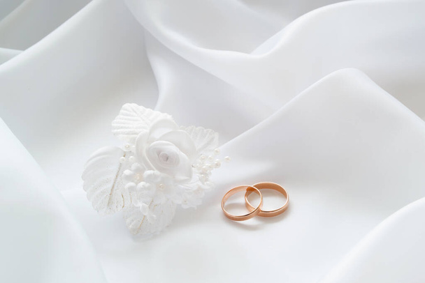 Gold wedding rings on white cloth and white artificial flowers to decorate the hairstyle. - Zdjęcie, obraz