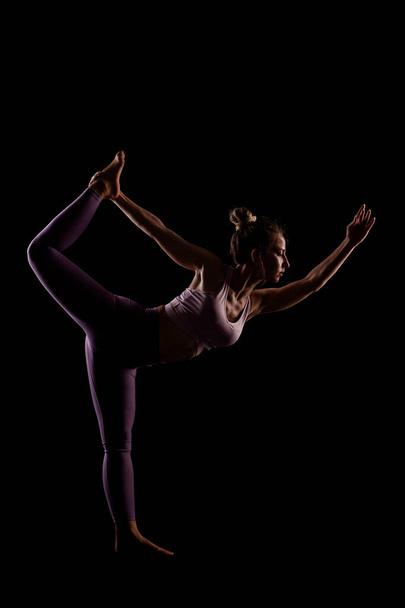 Fit girl practicing yoga in a studio. Half silhouette side lit fitness model. - Foto, afbeelding