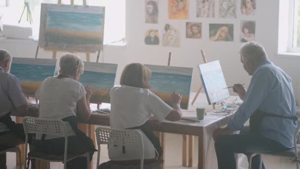Mladá učitelka výtvarného umění předvádí techniku malování akrylovými barvami na plátně pro skupinu důchodců. Skupina starších mužů a žen - Záběry, video