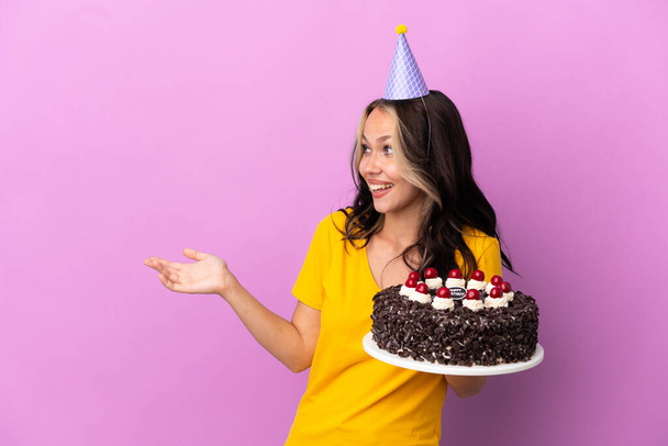 Adolescente chica rusa sosteniendo pastel de cumpleaños aislado sobre fondo púrpura con expresión sorpresa mientras mira al lado - Foto, imagen