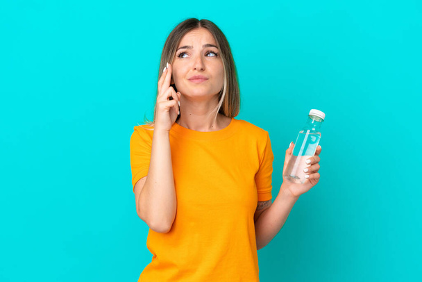 Young Romanian woman with a bottle of water isolated on blue background with fingers crossing and wishing the best - Foto, Imagem