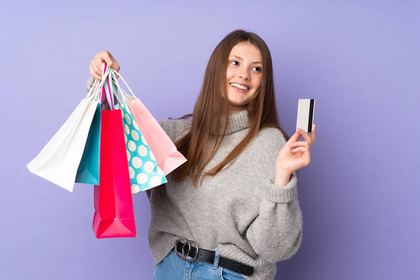 Adolescente caucásico chica aislado en púrpura fondo celebración de bolsas de compras y una tarjeta de crédito - Foto, imagen