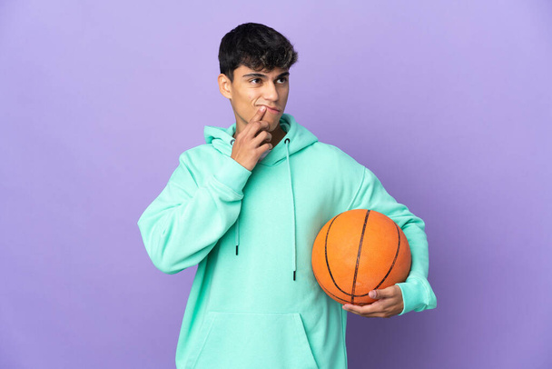 Jongeman basketbal spelen over geïsoleerde paarse achtergrond met twijfels tijdens het omhoog kijken - Foto, afbeelding