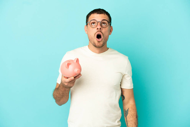 Brazilian man holding a piggybank over isolated blue background looking up and with surprised expression - Φωτογραφία, εικόνα