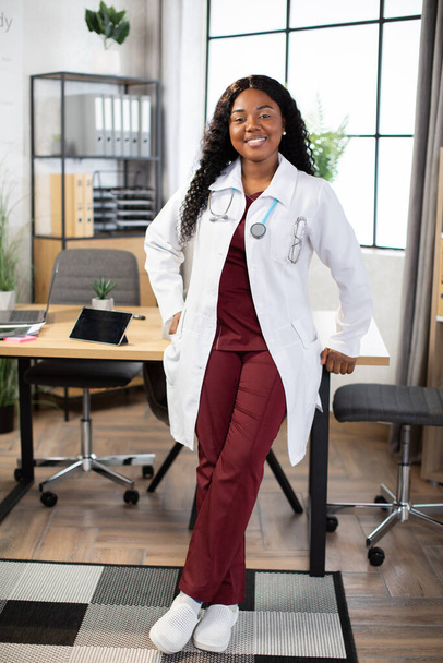 Pretty Afro-American woman doctor, posing to camera with happy smile in modern office - Foto, Bild