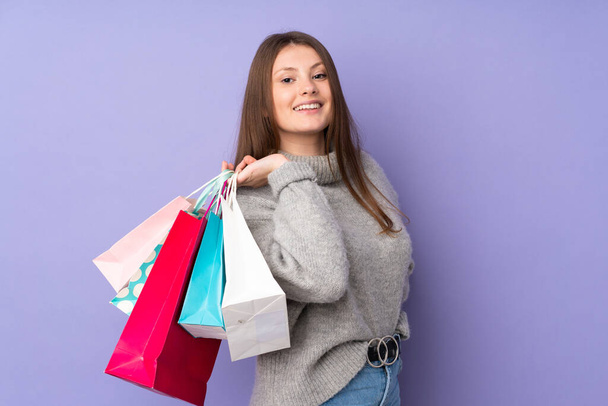 Adolescente caucásico chica aislado en púrpura fondo sosteniendo bolsas de compras y sonriendo - Foto, imagen