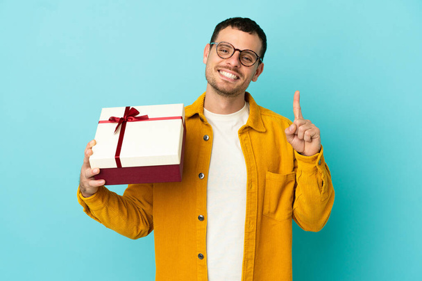 Brezilyalı bir adam mavi arka plan üzerinde bir hediye tutuyor ve en iyinin işareti olarak parmağını kaldırıyor. - Fotoğraf, Görsel