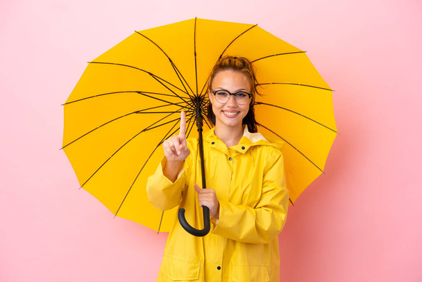 Tini orosz lány esőálló kabát és esernyő elszigetelt rózsaszín háttér mutatja, és emelje fel az ujját - Fotó, kép