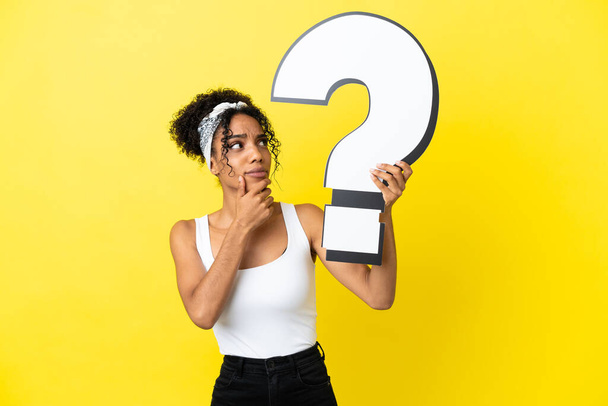 Fiatal afro-amerikai nő elszigetelt sárga háttér kezében kérdőjellel ikon és kétségei vannak - Fotó, kép