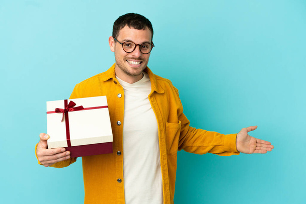 Brazilian man holding a gift over isolated blue background extending hands to the side for inviting to come - Photo, image