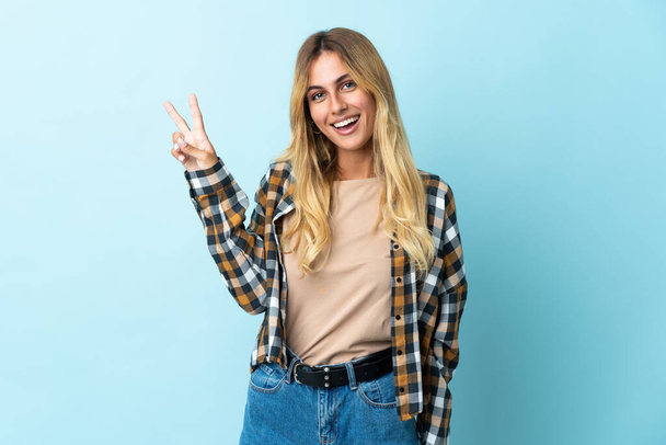 Junge blonde Uruguayerin auf blauem Hintergrund lächelt und zeigt Siegeszeichen - Foto, Bild