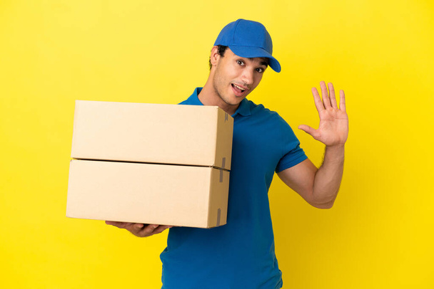 Delivery man over isolated yellow wall saluting with hand with happy expression - Photo, Image