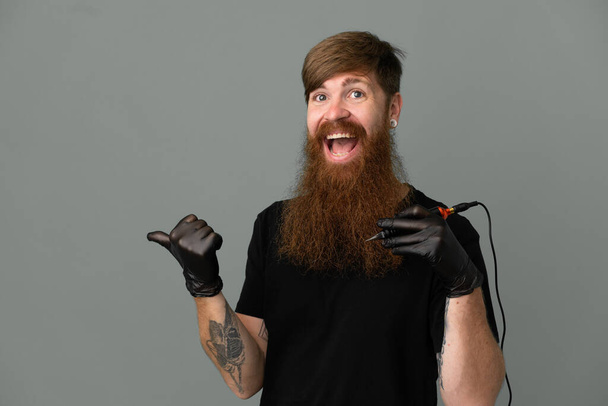 Tatuaggio artista rossa uomo isolato su sfondo blu che punta verso il lato per presentare un prodotto - Foto, immagini