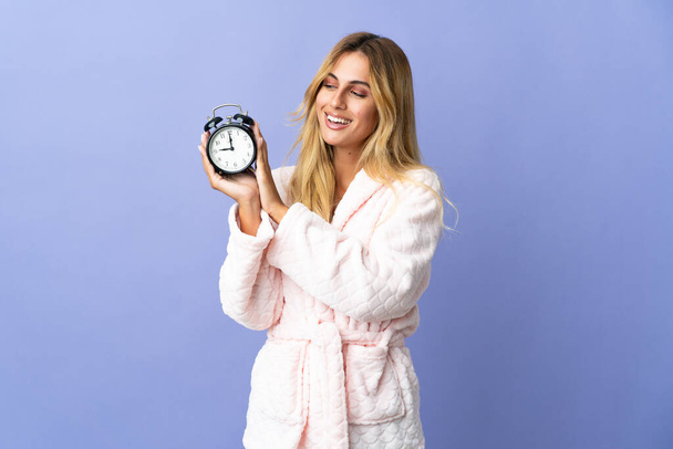 Young blonde Uruguayan woman isolated on blue background in pajamas and holding clock with happy expression - Φωτογραφία, εικόνα