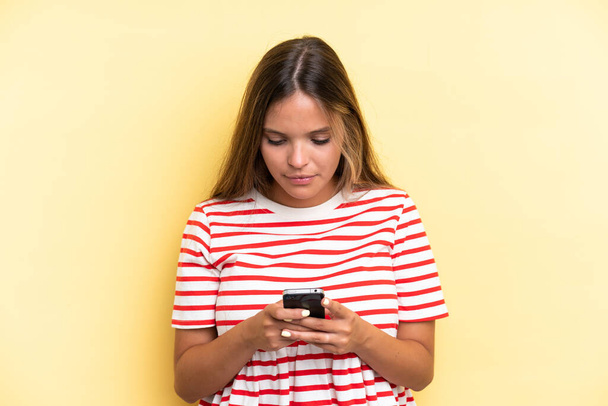 Young caucasian woman isolated on yellow background using mobile phone - Φωτογραφία, εικόνα