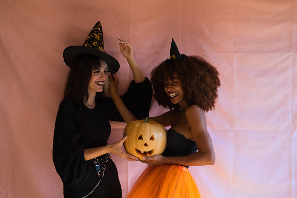 Две красивые молодые женщины, одна блондинка, а другая афроамериканка, одетые как ведьмы и держащие тыкву, веселятся на вечеринке в честь Хэллоуина. Женщины счастливы.. - Фото, изображение