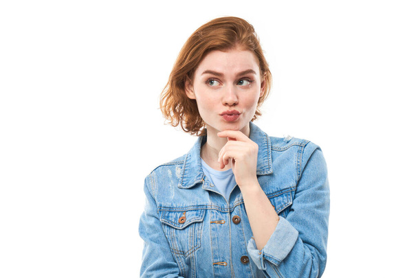 A girl student with red hair happily thinks and looks at the camera while holding her finger near her chin. Pensive thoughtful woman in a blue denim jacket on a white background. Copy space - Photo, Image
