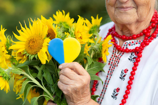 Κίτρινη μπλε καρδιά και ένα μπουκέτο ηλιοτρόπια στα χέρια μιας ηλικιωμένης γιαγιάς με κεντημένο πουκάμισο και κόκκινες χάντρες. Ημέρα ανεξαρτησίας της Ουκρανίας, σύνταγμα, σημαία - Φωτογραφία, εικόνα