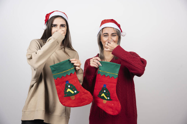 Két mosolygós lány télapó kalapban, karácsonyi zoknival, akik undorral zárják be az orrukat. Kiváló minőségű fénykép - Fotó, kép