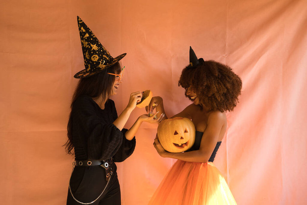 Młode Latynoski i Latynoski przebrane za czarownice śmieją się głośno i rozdają cukierki z dyni Halloween na różowym tle. - Zdjęcie, obraz