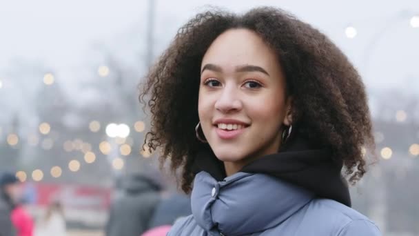 Side view of female portrait of African American girl with stylish afro hairstyle in curls stands outdoors on background of city skating rink and people, brunette looks at camera and smiles toothy - Footage, Video