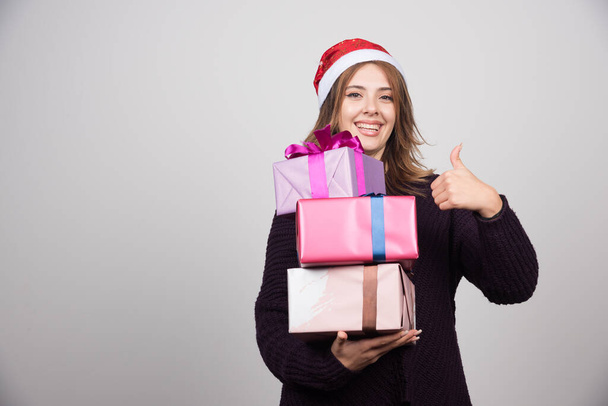 Young woman in Santa hat with gift boxes showing a thumb up. High quality photo - Фото, изображение