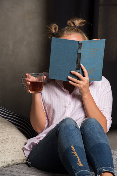 Frau verschließt ihr Gesicht mit Buch und hält heißen Tee auf dunklem Hintergrund  - Foto, Bild