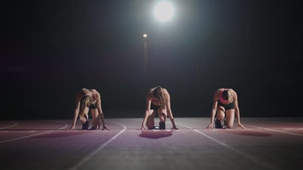 Fiatal, csinos nők kocognak sportruházatban és tornacipőben, együtt készülnek a futóversenyre a futópályán. Kezdje a sort, és indítsa el a versenyt lassított felvételen a sötétben - Felvétel, videó