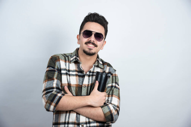 Photo du jeune homme dans des lunettes montrant un gobelet noir au-dessus d'un mur blanc. Photo de haute qualité - Photo, image