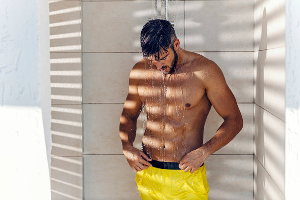El hombre se está duchando. Un hombre alto y guapo con músculos toma una ducha después de nadar. Un hombre semidesnudo en pantalones cortos de baño amarillos se para en la cabaña y se lava el pelo - Foto, imagen