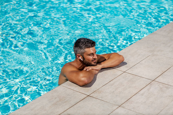 Sezon opalania i cieszenia się przy basenie. Poczuj słońce i muzykę przy basenie. Przystojny mężczyzna odświeża się w wodzie w basenie i słucha piosenki Odpoczywając i ciesząc się słońcem i latem - Zdjęcie, obraz