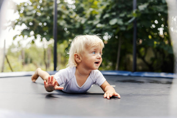 Liegen auf dem Trampolin nach dem Springen. Ein süßer kleiner Junge oder Mädchen in weißen Kinderkörpern spielt im Hof und springt auf einem Trampolin. Kleinkinder mit blonden Haaren und blauen Augen. Aufwachsen  - Foto, Bild