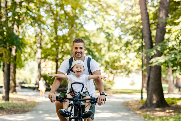 Father and son ride bicycles. Toddlers sit in a basket of bikes and wander around. Summer sunny family day, family bonding time. Active family weekend, vacation in the park with the family - Photo, Image
