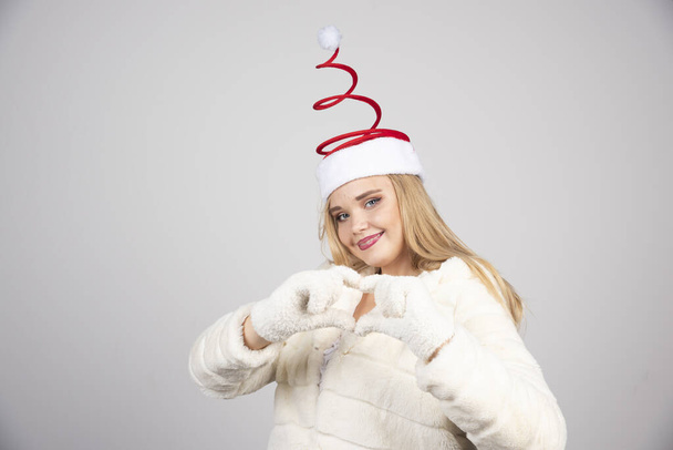 Nainen joulupukin hatussa tekemässä sydänkylttiä harmaalla taustalla. Laadukas kuva - Valokuva, kuva