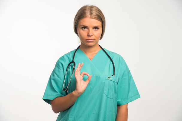 Blonde femme médecin en uniforme bleu avec stéthoscope dans le cou fait bon signe. Photo de haute qualité - Photo, image