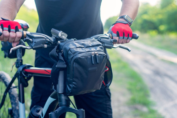 A cyclist in gloves near a mountain bike with a bag on the handlebars - Fotografie, Obrázek